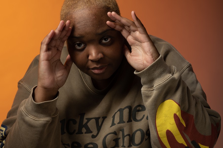 Rapper DijahSB is posed seated, with their hands on their head, with a warm backdrop behind them.