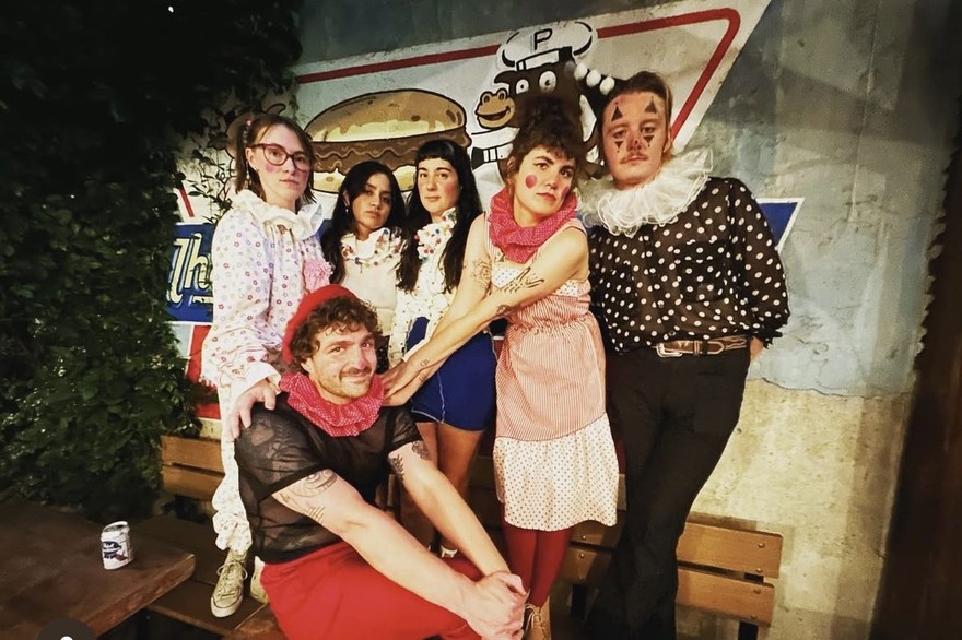 Six members of eclectic punk band Parisian Orgy are posed, standing on a bench while casually wearing clown attire