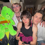 Photo taken indoors of eclectic four-piece Dial Up. One member is wearing a green teddy bear as a mask, another in bishop attire, one in black in pink striped hand warmers and the last with their hair spiked up.