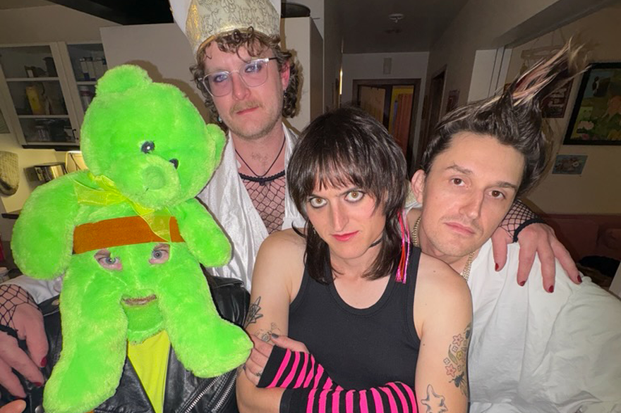 Photo taken indoors of eclectic four-piece Dial Up. One member is wearing a green teddy bear as a mask, another in bishop attire, one in black in pink striped hand warmers and the last with their hair spiked up.