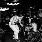 Black and white long exposure photo taken of the lead performer of Jed Arbour crouched toward an engaged crowd in a dimly lit venue.
