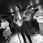 Black and white photo taken of Bugswallow performing on stage at the Palomino. Front person is singing and playing steel guitar, image also contains a member playing banjo and one on keyboard.