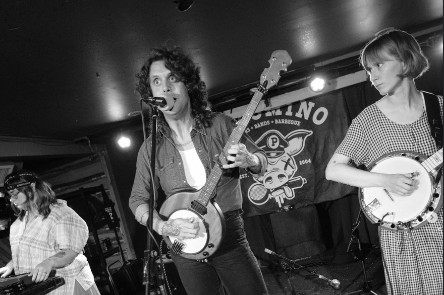 Black and white photo taken of Bugswallow performing on stage at the Palomino. Front person is singing and playing steel guitar, image also contains a member playing banjo and one on keyboard.