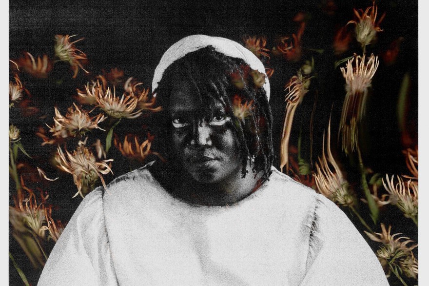 Solo artist Backxwash is photographed in black and white. Warm toned flowers fill the background.