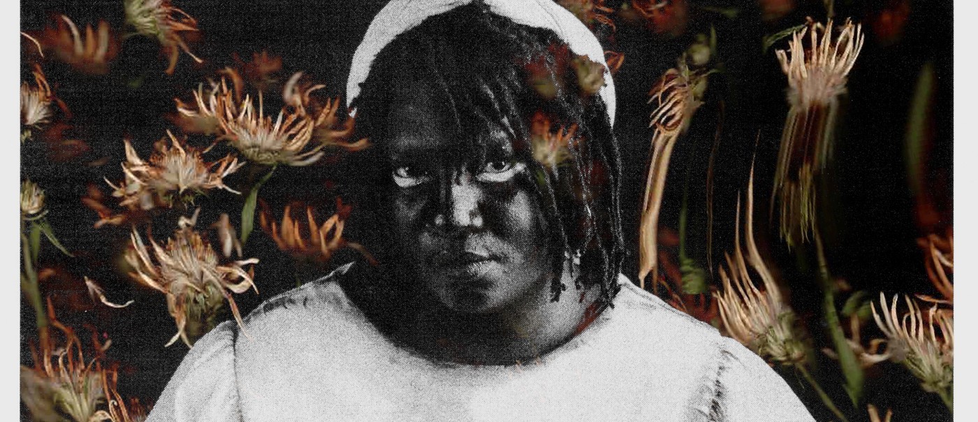 Solo artist Backxwash is photographed in black and white. Warm toned flowers fill the background.