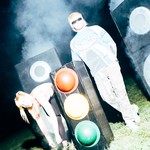 Two members of Snõõper stand outdoors at night in heavy artificial fog, surrounded by large speaker‑like boxes and a traffic light. One person in sunglasses and a blue‑and‑white long‑sleeve shirt stands upright, while the other, wearing a pink‑and‑white top, bends forward near the traffic light. Bright flash lighting creates a stark, surreal scene against the dark background.