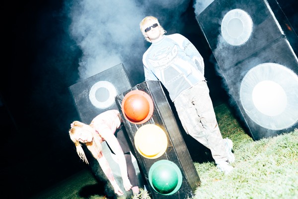 Two members of Snõõper stand outdoors at night in heavy artificial fog, surrounded by large speaker‑like boxes and a traffic light. One person in sunglasses and a blue‑and‑white long‑sleeve shirt stands upright, while the other, wearing a pink‑and‑white top, bends forward near the traffic light. Bright flash lighting creates a stark, surreal scene against the dark background.