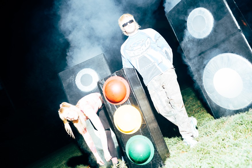Two members of Snõõper stand outdoors at night in heavy artificial fog, surrounded by large speaker‑like boxes and a traffic light. One person in sunglasses and a blue‑and‑white long‑sleeve shirt stands upright, while the other, wearing a pink‑and‑white top, bends forward near the traffic light. Bright flash lighting creates a stark, surreal scene against the dark background.