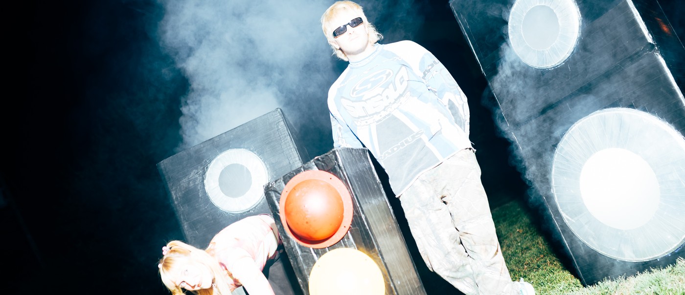 Two members of Snõõper stand outdoors at night in heavy artificial fog, surrounded by large speaker‑like boxes and a traffic light. One person in sunglasses and a blue‑and‑white long‑sleeve shirt stands upright, while the other, wearing a pink‑and‑white top, bends forward near the traffic light. Bright flash lighting creates a stark, surreal scene against the dark background.