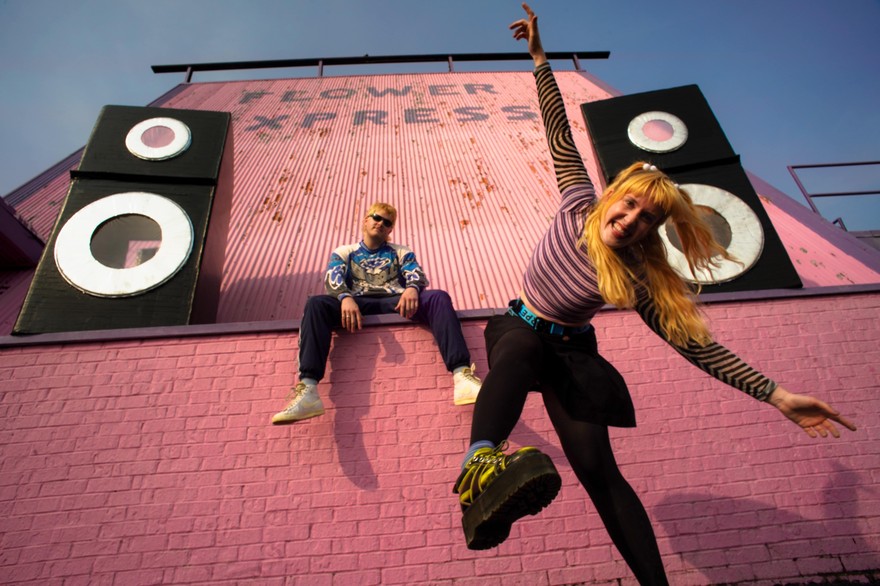 Two members of Snõõper are posed eclectically in front of pink brick, with two large speakers on each side.