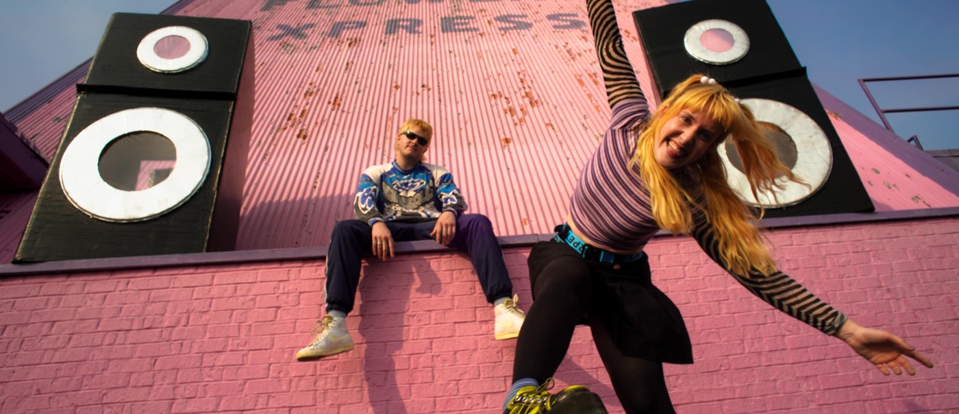 Two members of Snõõper are posed eclectically in front of pink brick, with two large speakers on each side.