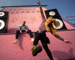 Two members of Snõõper are posed eclectically in front of pink brick, with two large speakers on each side.