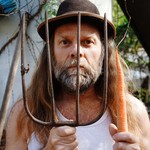 Thor Harris stands behind the tines of a pitchfork with a carrot hanging from one tine, looking through the metal prongs with a calm, focused expression.