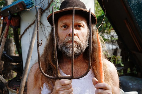 Thor Harris stands behind the tines of a pitchfork with a carrot hanging from one tine, looking through the metal prongs with a calm, focused expression.