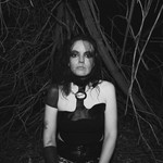 Black and white photo of experimental artist Lana del Rabies, dressed in black, staring into the camera in a forested setting.