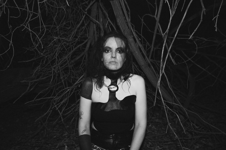 Black and white photo of experimental artist Lana del Rabies, dressed in black, staring into the camera in a forested setting.
