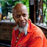 Photo of solo artist Laraaji, posed facing the camera wearing an orange coat.