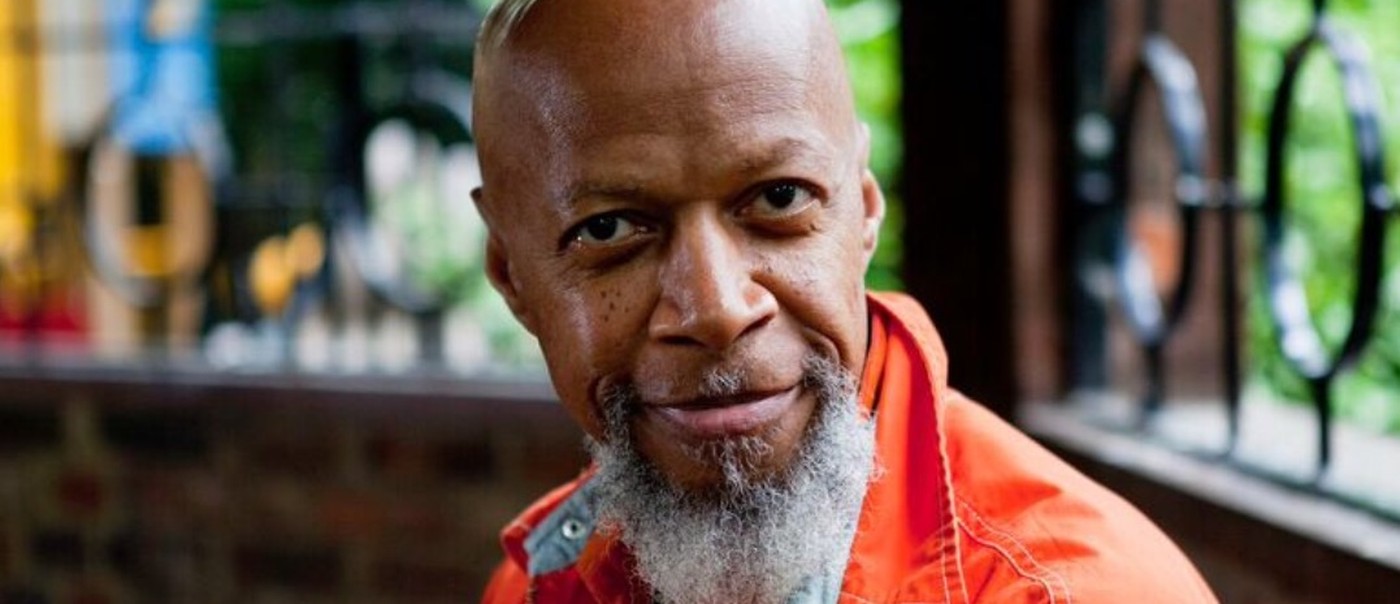 Photo of solo artist Laraaji, posed facing the camera wearing an orange coat.
