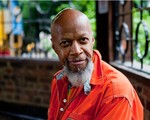 Photo of solo artist Laraaji, posed facing the camera wearing an orange coat.