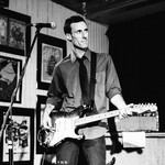 Black‑and‑white photo of musician Chris Burns performing on a small stage. He stands at a microphone with an electric guitar, wearing a collared shirt with rolled‑up sleeves and a tie. Framed posters and artwork fill the wall behind him, and an amplifier sits to his right. The scene has an intimate, vintage live‑show atmosphere.