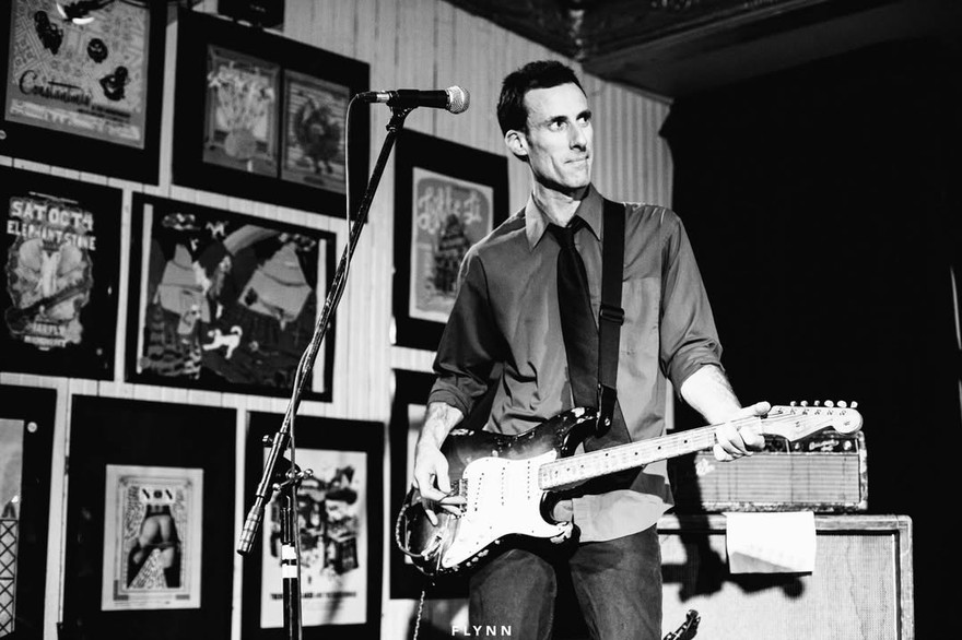 Black‑and‑white photo of musician Chris Burns performing on a small stage. He stands at a microphone with an electric guitar, wearing a collared shirt with rolled‑up sleeves and a tie. Framed posters and artwork fill the wall behind him, and an amplifier sits to his right. The scene has an intimate, vintage live‑show atmosphere.