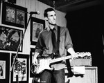Black‑and‑white photo of musician Chris Burns performing on a small stage. He stands at a microphone with an electric guitar, wearing a collared shirt with rolled‑up sleeves and a tie. Framed posters and artwork fill the wall behind him, and an amplifier sits to his right. The scene has an intimate, vintage live‑show atmosphere.
