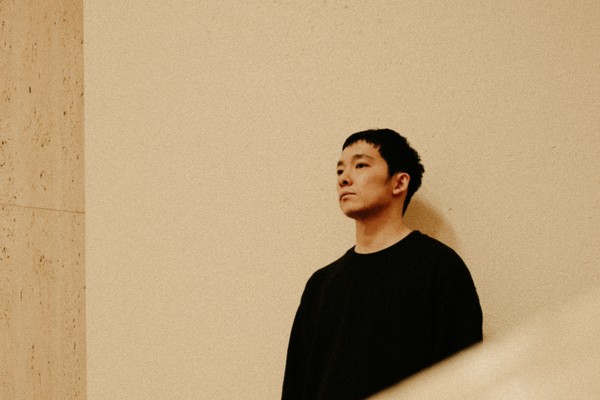 Photo of Mineo Kawasaki wearing all black, posed looking away from the camera as he stands in front of a neutral wall.