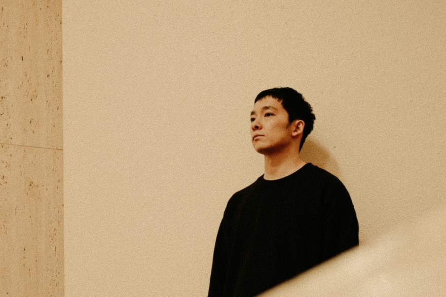 Photo of Mineo Kawasaki wearing all black, posed looking away from the camera as he stands in front of a neutral wall.