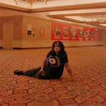 Lead vocalist of Palehound, El Kempner is photographed, on a patterned carpeted floor in a large indoor space, wearing a black tie‑dye shirt with a white eight‑ball‑like design, ripped dark pants, and black shoes. Warm lighting softens the beige and red walls behind them, which are covered with posters and ads, creating a quiet, candid moment in a public hallway.