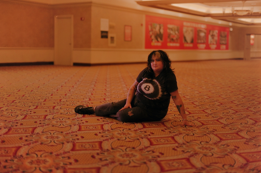 Lead vocalist of Palehound, El Kempner is photographed, on a patterned carpeted floor in a large indoor space, wearing a black tie‑dye shirt with a white eight‑ball‑like design, ripped dark pants, and black shoes. Warm lighting softens the beige and red walls behind them, which are covered with posters and ads, creating a quiet, candid moment in a public hallway.