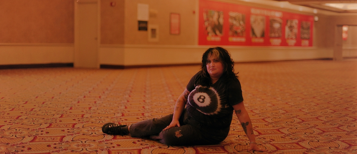 Lead vocalist of Palehound, El Kempner is photographed, on a patterned carpeted floor in a large indoor space, wearing a black tie‑dye shirt with a white eight‑ball‑like design, ripped dark pants, and black shoes. Warm lighting softens the beige and red walls behind them, which are covered with posters and ads, creating a quiet, candid moment in a public hallway.