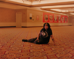Lead vocalist of Palehound, El Kempner is photographed, on a patterned carpeted floor in a large indoor space, wearing a black tie‑dye shirt with a white eight‑ball‑like design, ripped dark pants, and black shoes. Warm lighting softens the beige and red walls behind them, which are covered with posters and ads, creating a quiet, candid moment in a public hallway.