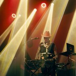 Christopher Fleeger of Cooling Prongs captured onstage, performing on a keyboard, bathed in yellow and red lighting, wearing a bright cowboy hat and boots.