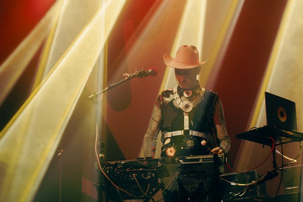 Christopher Fleeger of Cooling Prongs captured onstage, performing on a keyboard, bathed in yellow and red lighting, wearing a bright cowboy hat and boots.