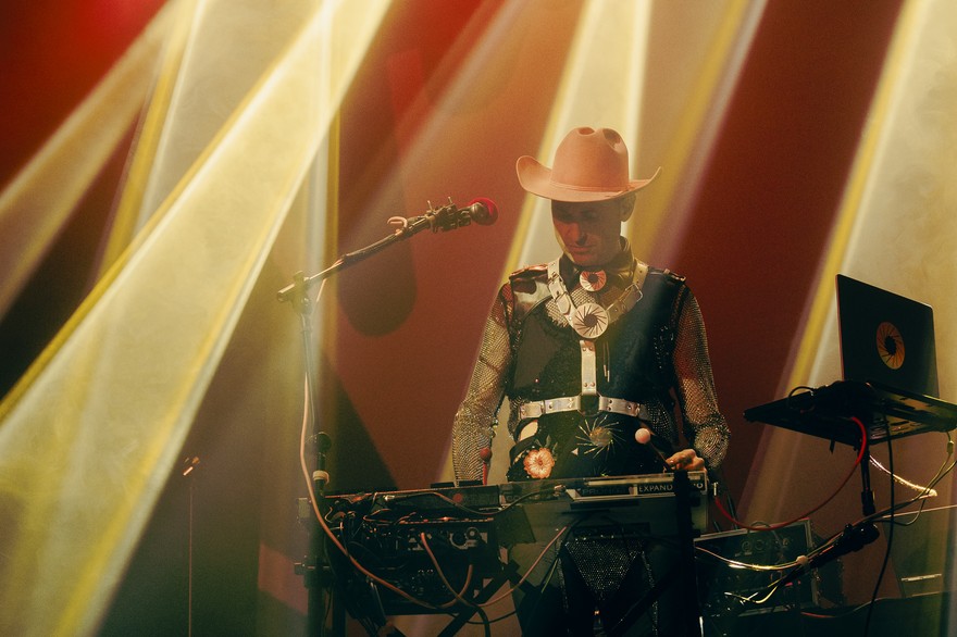 Christopher Fleeger of Cooling Prongs captured onstage, performing on a keyboard, bathed in yellow and red lighting, wearing a bright cowboy hat and boots.