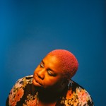 Portrait of Sharon Udoh with bright pink hair against a solid blue background, wearing a floral garment with large pink, orange, and cream flowers. Warm lighting highlights her face and hair, creating a vivid contrast with the cool backdrop.