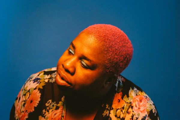 Portrait of Sharon Udoh with bright pink hair against a solid blue background, wearing a floral garment with large pink, orange, and cream flowers. Warm lighting highlights her face and hair, creating a vivid contrast with the cool backdrop.