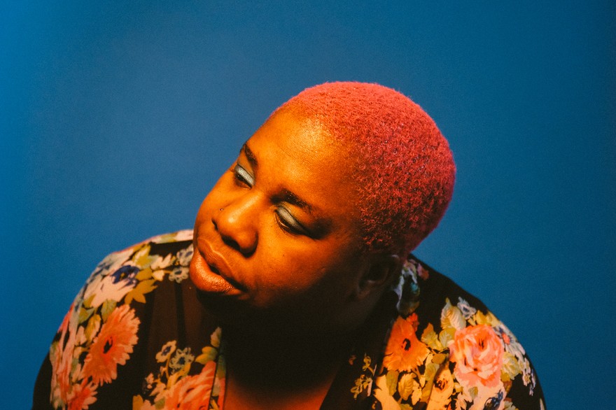 Portrait of Sharon Udoh with bright pink hair against a solid blue background, wearing a floral garment with large pink, orange, and cream flowers. Warm lighting highlights her face and hair, creating a vivid contrast with the cool backdrop.