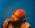 Portrait of Sharon Udoh with bright pink hair against a solid blue background, wearing a floral garment with large pink, orange, and cream flowers. Warm lighting highlights her face and hair, creating a vivid contrast with the cool backdrop.