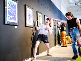 Two people interact playfully inside an art‑gallery space at Sled Village 2025. One person leans back with arms raised while another points toward them in an exaggerated gesture. Framed posters with bold, colorful designs line the dark wall behind them, and another attendee looks at the artwork in the background.