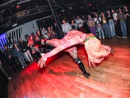 Drag Queen Dayna Hart performs at the Sled Island 2025 kickoff at Commonwealth, captured mid‑movement in a dramatic back‑arched pose on a wooden floor. She wears a pink patterned outfit and high black boots, surrounded by a semicircle of onlookers watching closely in the dim, club‑like venue.