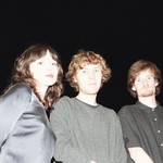 Three members of Flooding stand close together in front of a pitch‑black background, illuminated by a bright flash. One wears a shiny satin long‑sleeve top, another a textured gray sweater, and the third a short‑sleeve black button‑up shirt. Their faces and upper bodies are sharply lit while everything behind them fades into darkness.