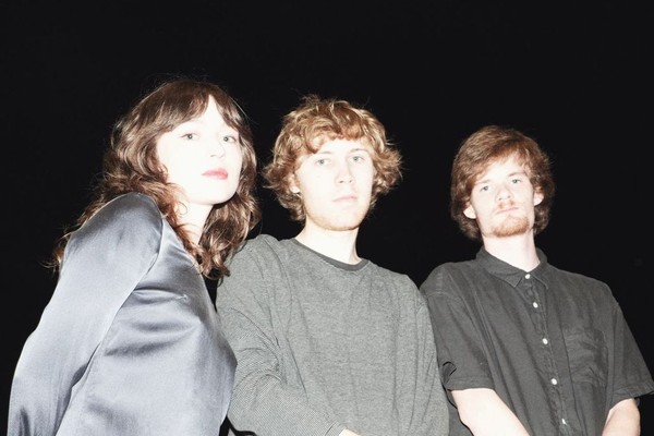 Three members of Flooding stand close together in front of a pitch‑black background, illuminated by a bright flash. One wears a shiny satin long‑sleeve top, another a textured gray sweater, and the third a short‑sleeve black button‑up shirt. Their faces and upper bodies are sharply lit while everything behind them fades into darkness.
