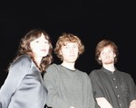 Three members of Flooding stand close together in front of a pitch‑black background, illuminated by a bright flash. One wears a shiny satin long‑sleeve top, another a textured gray sweater, and the third a short‑sleeve black button‑up shirt. Their faces and upper bodies are sharply lit while everything behind them fades into darkness.