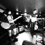 Black‑and‑white photo of Spec Realists performing onstage at the Palomino. Three musicians play electric guitars under bright stage lights, with a drum kit behind them and a “Palace” banner hanging in the background. The scene captures the energy of an intimate live show.