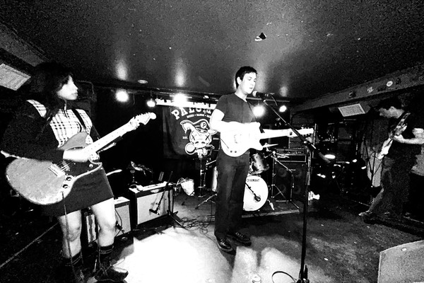 Black‑and‑white photo of Spec Realists performing onstage at the Palomino. Three musicians play electric guitars under bright stage lights, with a drum kit behind them and a “Palace” banner hanging in the background. The scene captures the energy of an intimate live show.