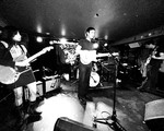 Black‑and‑white photo of Spec Realists performing onstage at the Palomino. Three musicians play electric guitars under bright stage lights, with a drum kit behind them and a “Palace” banner hanging in the background. The scene captures the energy of an intimate live show.