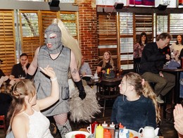 A drag performer in a gray plaid outfit, fishnet stockings, long black gloves, and a full head mask with a long blonde ponytail leans toward a seated guest during a lively drag brunch at the Palomino for Sled Island 2025. The table in front is crowded with food, red cups, and drinks, while another person photographs the moment and diners in the background watch the interaction.