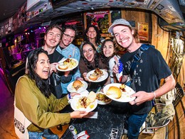 Seven people stand together inside the Palomino during Sled Island 2026’s pig roast, each holding plates piled with burgers, sandwiches, and sides. Behind them, a small stage with instruments, colorful lights, and poster‑covered walls creates a lively, casual bar atmosphere.