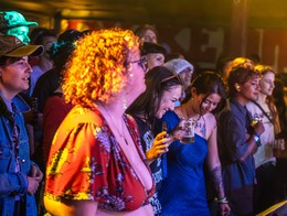 A crowd gathers inside the Palomino during a Sled Island 2025 show, lit by shifting yellow, blue, and green stage lights. People stand shoulder to shoulder, some holding drinks, all facing the performance with focused, excited energy in the dim, colorful venue.